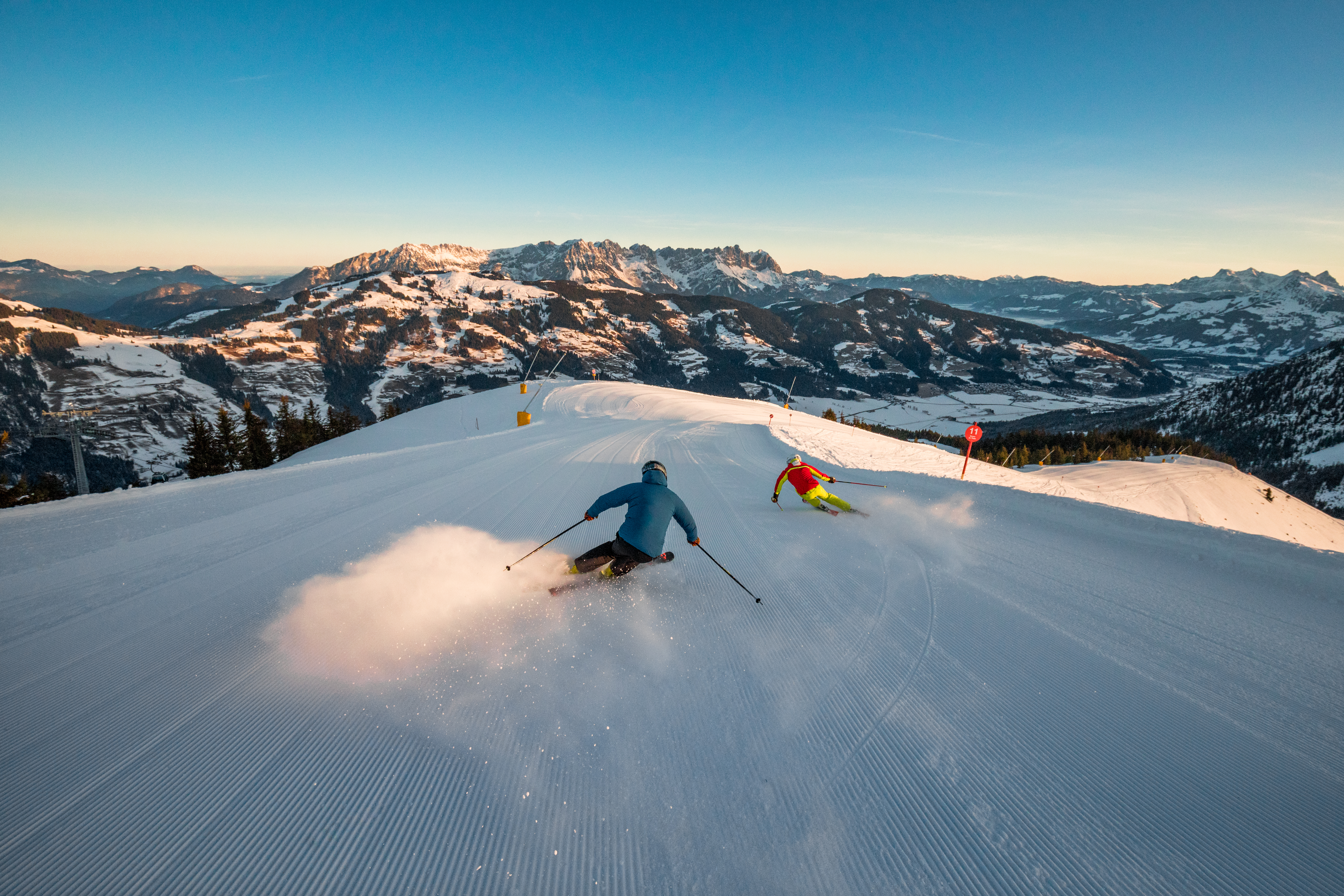 Skiregion - Tirol - Zwei Skifahrer gleiten die schneebedeckte Piste hinunter, umgeben von majestätischen Bergen. Der Himmel ist klar und blau, die Landschaft strahlt winterliche Ruhe aus. - SkiWelt Wilder Kaiser - Brixental